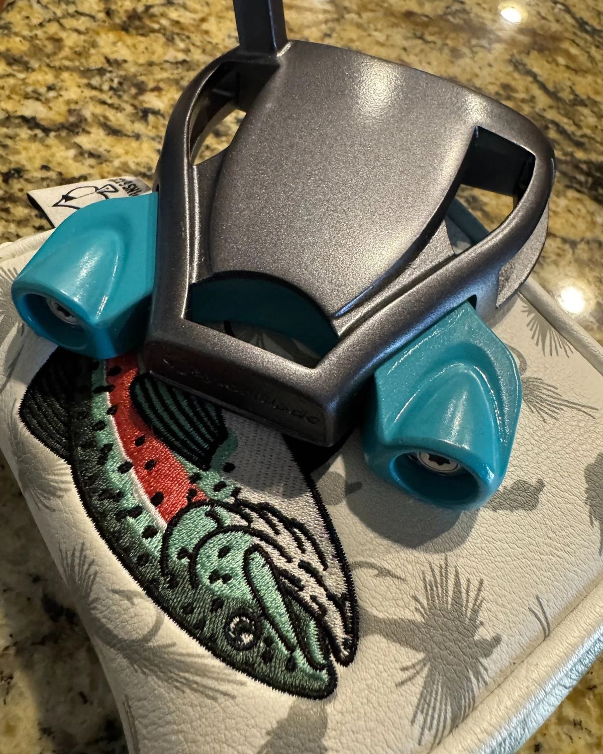 Teal and silver mallet putter with custom paint fill, resting on a trout-embroidered headcover